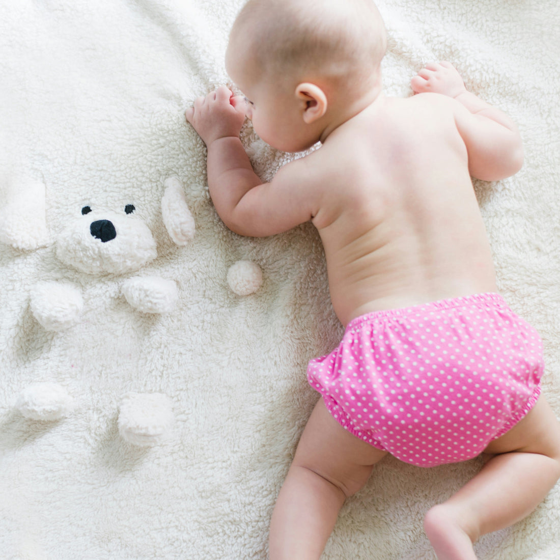 A baby wearing pink pants and crawling besides a toy bear